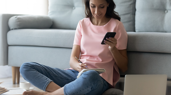 Junge Frau sitzt auf dem Boden vor einem Sofa und schaut auf ihr Smartphone, das sie in der Hand hält.