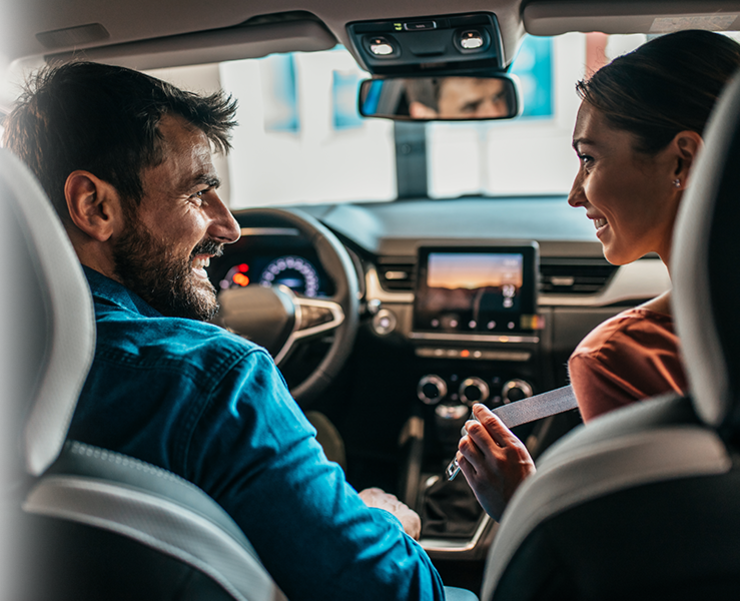 Ein Mann und eine Frau, die vorne in einem Auto sitzen, sich anschauen und anlächeln.