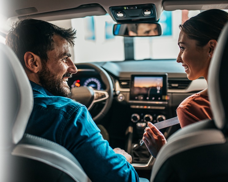 Ein Mann und eine Frau, die vorne in einem Auto sitzen, sich anschauen und anlächeln.