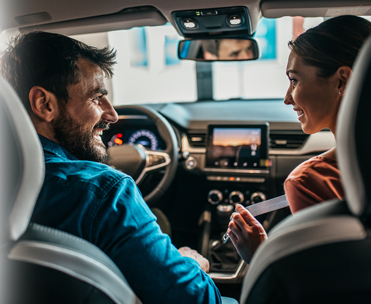 Ein Mann und eine Frau, die vorne in einem Auto sitzen, sich anschauen und anlächeln.