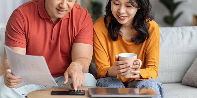 Ein Mann und eine Frau sitzen auf dem Sofa, vor ihnen auf dem Couchtisch liegt ein Taschenrechner. Der Mann tippt etwas in den Taschenrechner ein, beide blicken auf den Taschenrechner.