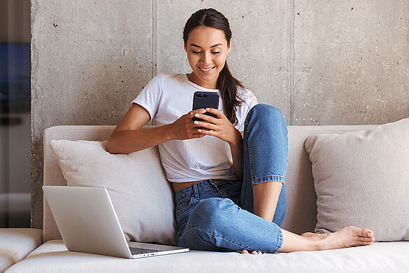 Frau sitzt mit Laptop und Smartphone auf Sofa