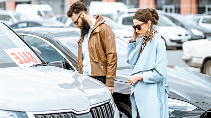 Ein Mann und eine Frau, die sich auf dem Außengelände eines Autohauses befinden. Um sie herum stehen einige zu verkaufende Autos, die sich die beiden anschauen.